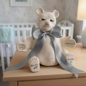Beige ceramic teddy with grey ribbon sat on a chest of drawers in front of a babys crib. 